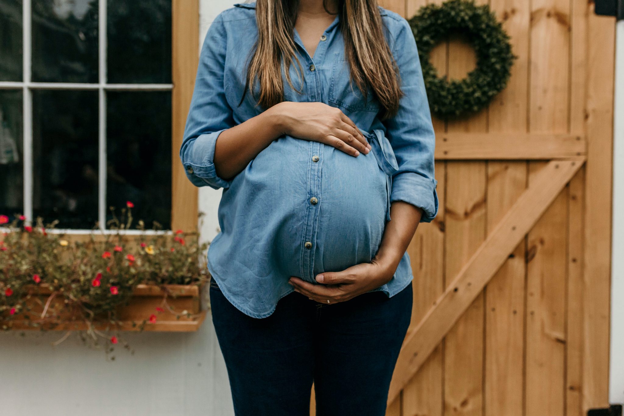 Birth Baby woman in blue denim button up jacket and blue denim jeans standing near brown wooden fence
