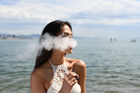Vaping woman wearing white sleeveless top smoking tobacco while standing near blue sea under white and blue skies during daytime