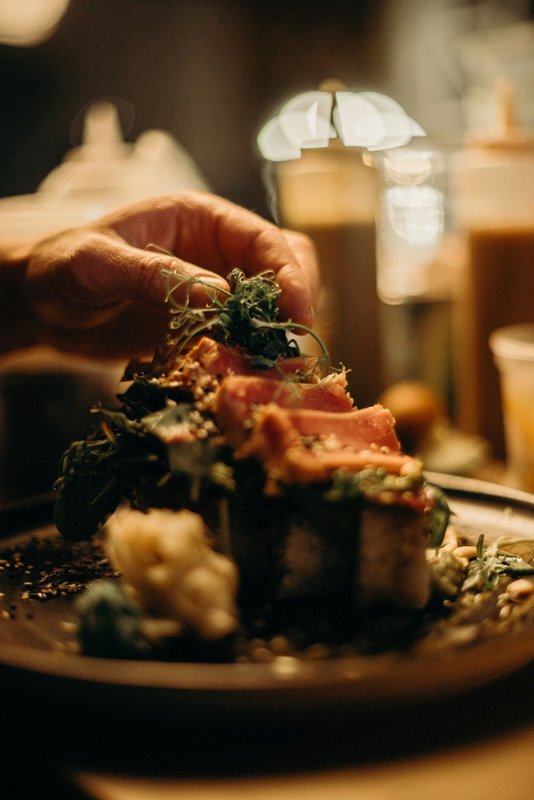 A cook garnishing a plated fish dish with herbs in a dim restaurant